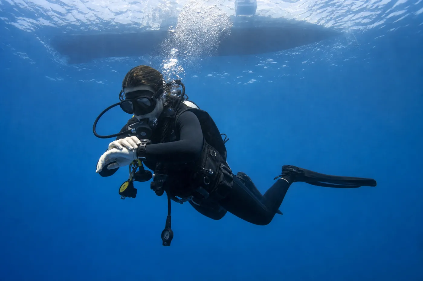 Diver holding a controlled five metre safety stop in blue water while checking a dive computer, exhaled bubbles rising in a steady stream