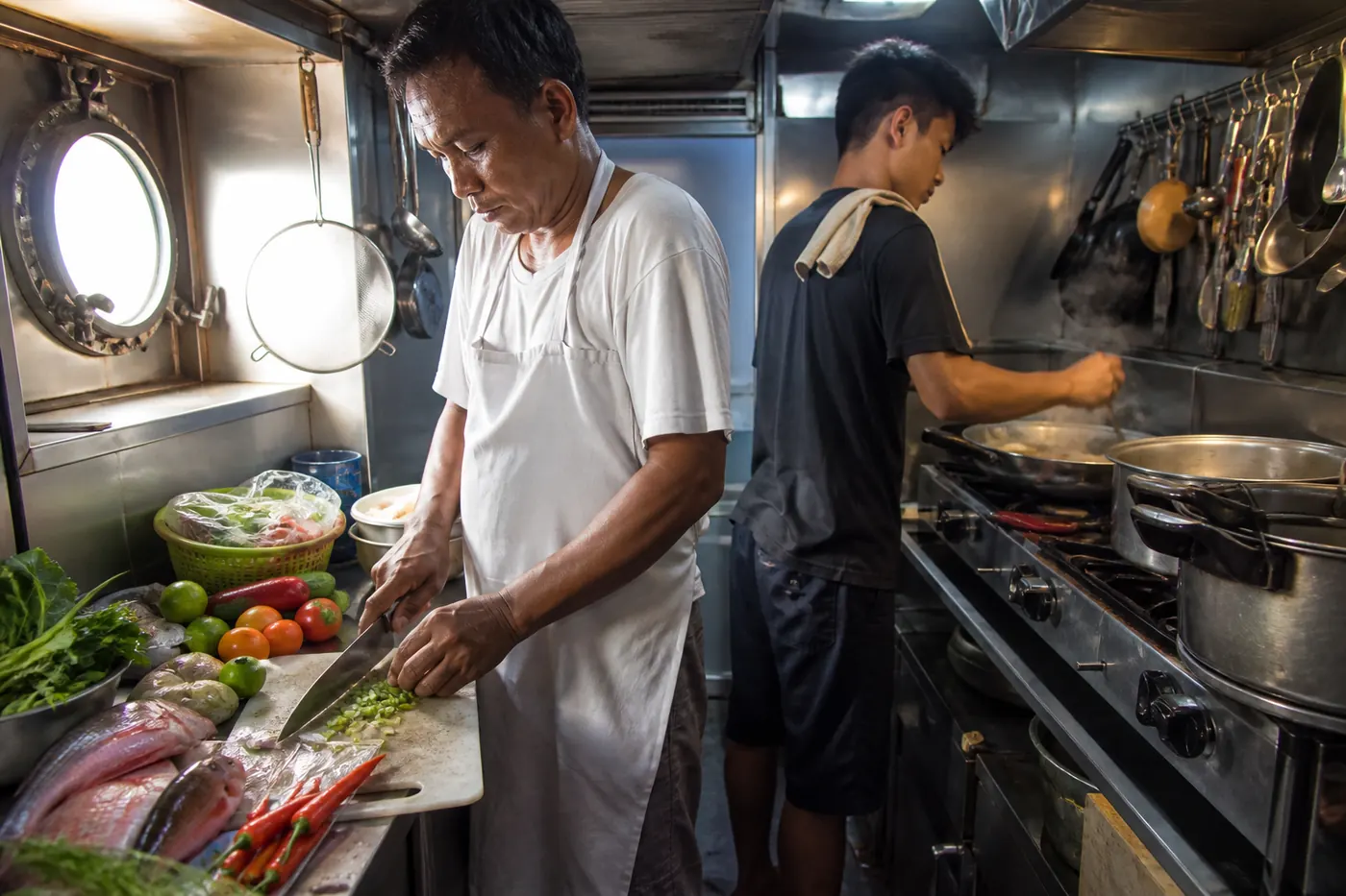 An Indonesian liveaboard chef and galley assistant preparing a meal in the compact stainless steel kitchen below deck, chopping vegetables and tending pans, working out of sight of the guests who will eat four meals a day on the trip