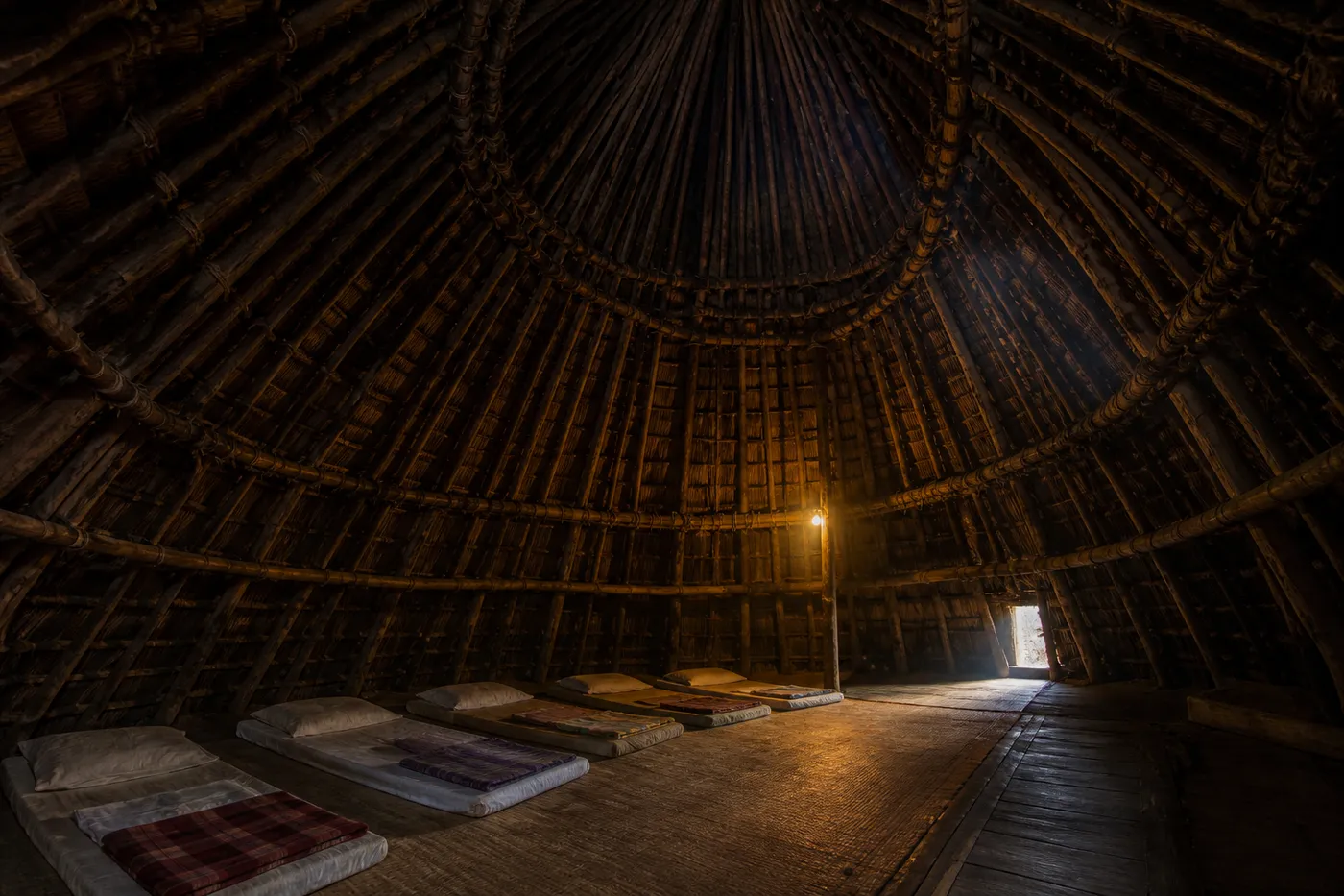 Interior of a mbaru niang house at Wae Rebo, Flores, showing the timber frame and palm thatch from inside, woven floor mats and bedding laid out in low evening light