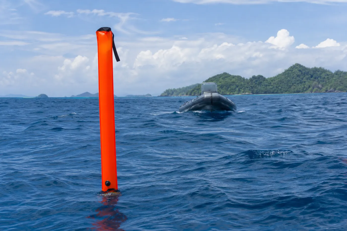 A tall orange surface marker buoy floating upright on choppy open water in Indonesia, with a small dive tender approaching in the distance under a bright sky