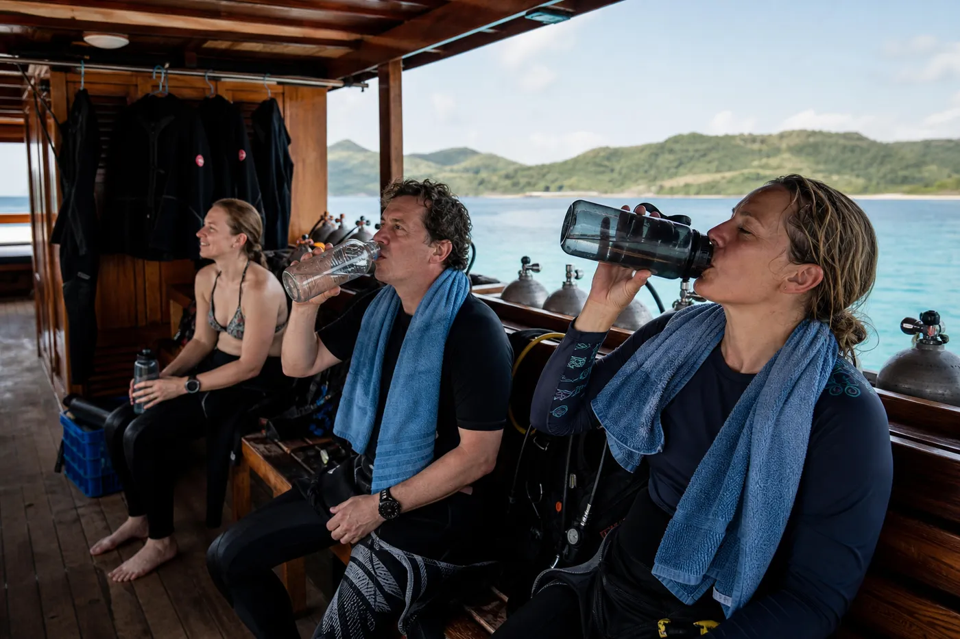 Divers resting on the shaded dive deck of a liveaboard between dives, towels over shoulders and water bottles alongside the tank racks