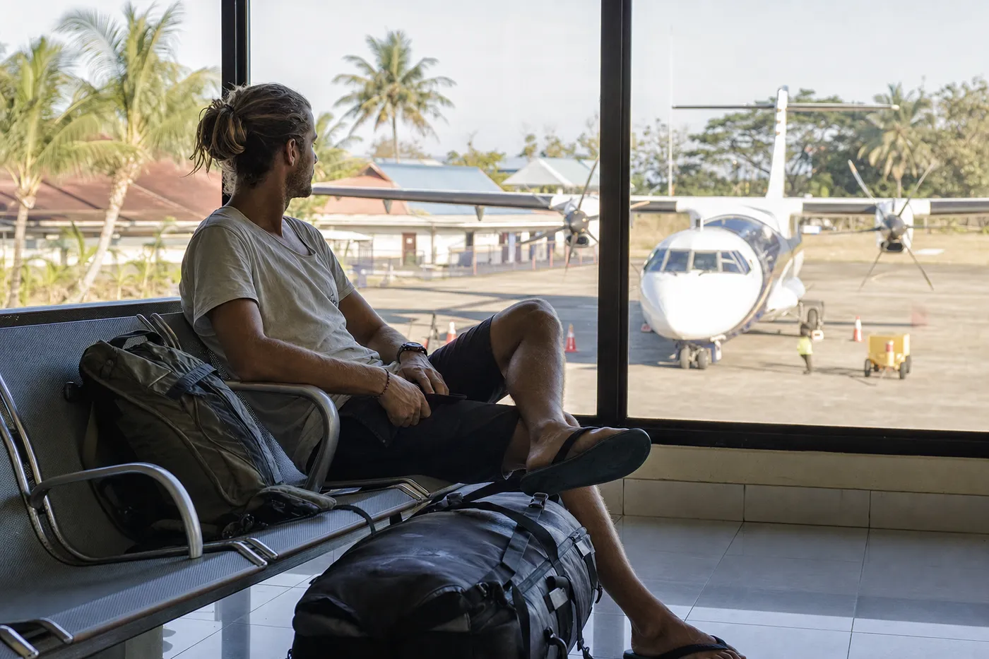 Traveler at a small Indonesian airport gate with a dive gear bag, looking at a departures board, morning light, relaxed after a liveaboard trip