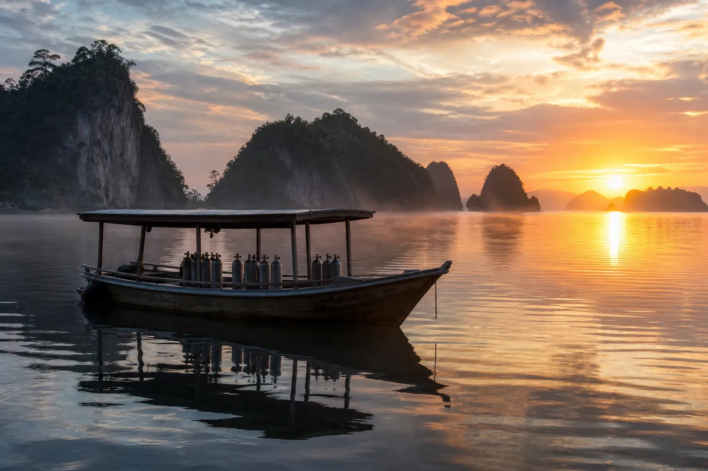 A dive boat on flat calm water off Kri Island in Raja Ampat at sunrise, with low golden light on forested limestone islands and mist on the water before the first dive