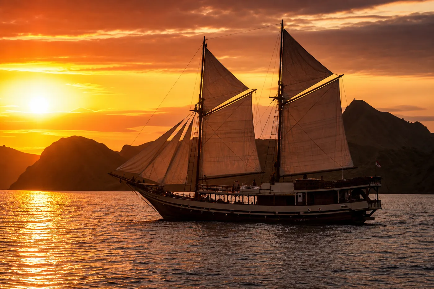 Traditional two-masted phinisi sailing yacht with sails set at sunset near Padar Island in Komodo National Park, golden light on the water